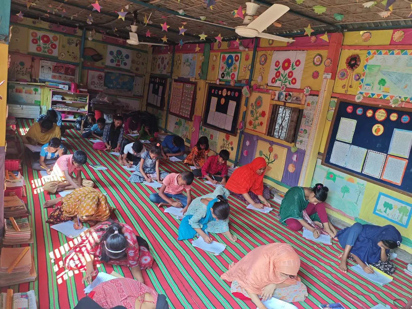 A full BRAC classroom in Bangladesh with students sitting on striped mats, writing in their notebooks