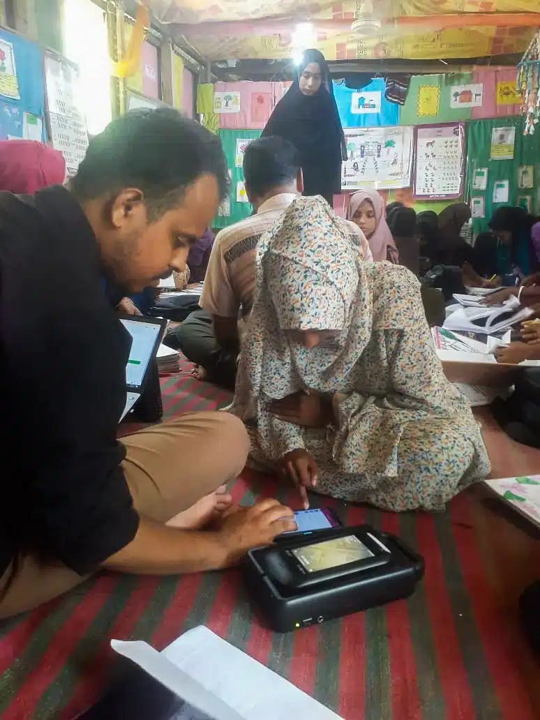 BRAC staff and a teacher recording attendance on a portable Tipsoi device on a classroom floor