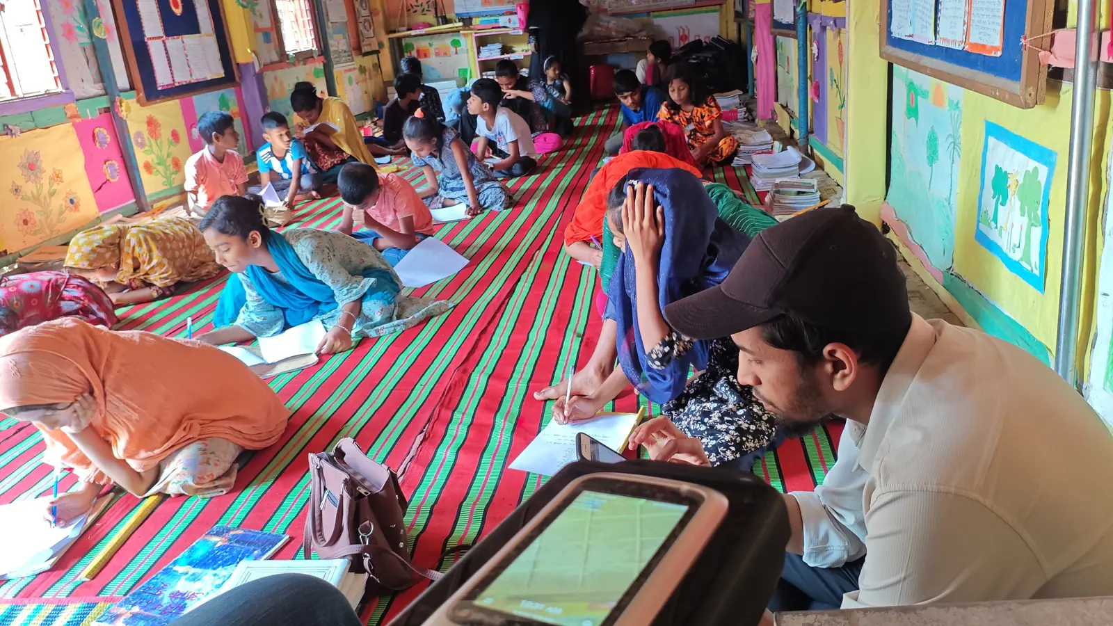 BRAC school classroom in rural Bangladesh where students sit on a mat writing, with a Tipsoi attendance device in the foreground