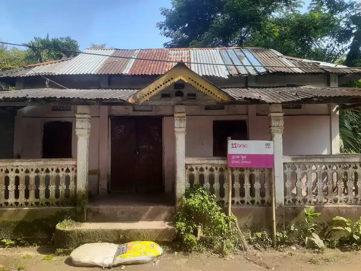 A rural BRAC school building in Bangladesh with a tin roof and a BRAC School sign by the veranda